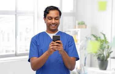 medicine, healthcare and technology concept - happy smiling indian doctor or male nurse in blue uniform using smartphone over medical office at hospital background