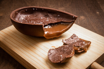 Cracked chocolate easter egg with flower of salt, stuffed with dulce de leche (dripping) on a wooden stand on a wooden table.