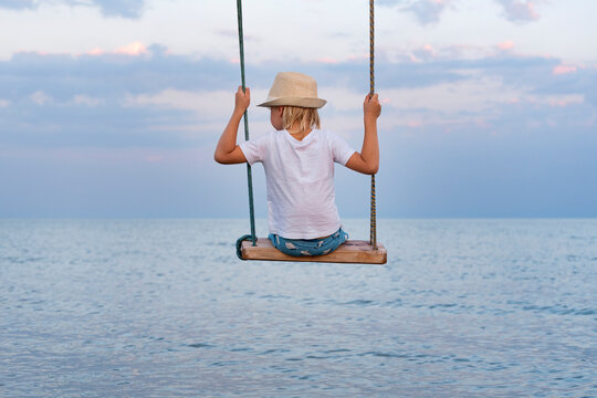 Child Sits On Swing Above The Water And Looks Out To Sea. Happy Childhood. The Rest Of The Sea.