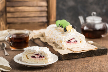 Meringue roll with berry filling with a cut piece on a plate, with a teapot and a mug of tea on a dark background.