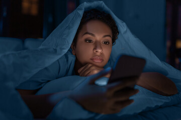 technology, internet, communication and people concept - young african american woman with smartphone lying under blanket in bed at home at night