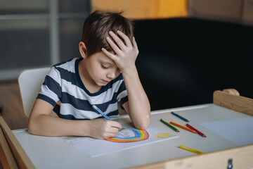 adorable caucasian boy of elementary age drawing a rainbow with pencils sitting at the desk in his room with head reclined upon his hand. Image with selective focus