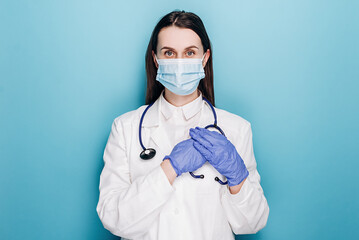 Dreamy young female nurse or doctor in protective face mask and latex gloves, press hands to heart, daydreaming, keep in mind, isolated on blue background. Medical workers, covid-19 pandemic concept