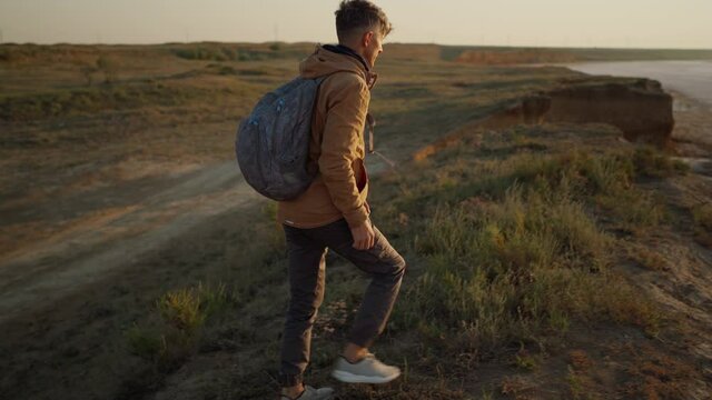 side view man hiker in casual wear climbs on steep grassy hill sea coast and looking sunrise at deserted wide sea beach, enjoying freedom, fresh air and nature beauty