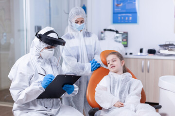 Orthodontist specialist writing treatment dressed in coverall in the course of coronavirus outbreak and kid consultation. Stomatologist during covid19 wearing ppe suit doing teeth procedure of child