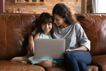 Happy young Latino mother and small daughter relax on couch at home watch video on laptop together....