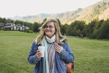 Young blonde woman in jeans coat with paper cup of morning coffee at the chalet cottage village on...