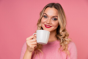 Drink coffee, enjoy a hot drink. A young woman in a warm pink sweater on a pink background with a white mug in her hands.