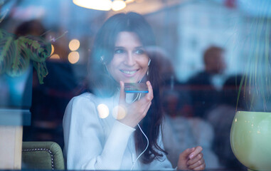 Shot through glass, a beautiful young European woman with headphones and a phone in her hands...