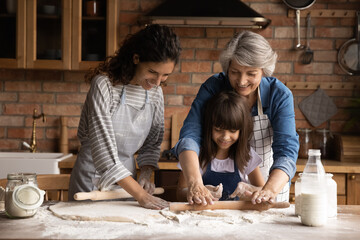 Smiling three generations of Hispanic women have fun baking together with dough at home kitchen....