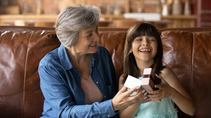 Smiling mature Hispanic grandmother give present to excited little girl child congratulate with birthday. Happy senior grandparent greeting small granddaughter with gift in box. Celebration concept.