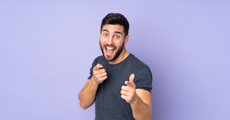 Caucasian handsome man surprised and pointing front over isolated purple background