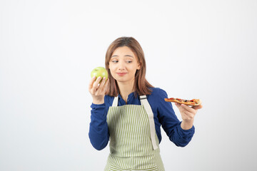 Portrait of woman in apron trying to choose what to eat apple or pizza