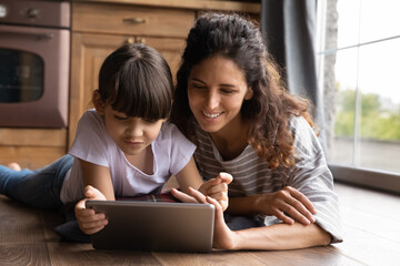 Caring young Hispanic mom and little daughter use modern tablet watch video online together. Happy...