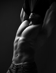 Black and white portrait of handsome muscular man, athlete with perfect built body standing taking off his shirt, getting naked, demonstrating perfect abs and chest muscles over dark background