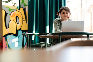 Smiling girl using headphones and laptop while sitting in cafe