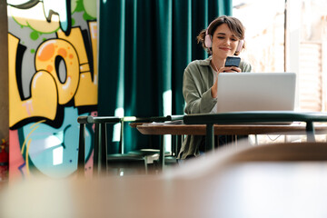 Girl in headphones using cellphone and laptop while sitting in cafe