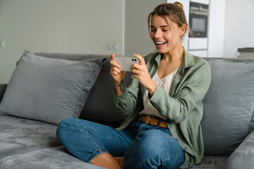 Happy woman playing online game on mobile phone while sitting on sofa