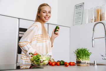 Obraz na płótnie Canvas Joyful blonde woman holding cucumber while preparing fresh salad