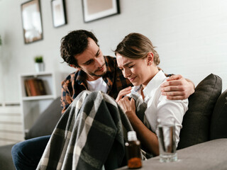 Young woman sick in bed. .Woman drinking medicine, having stomach pain. Husband and wife sitting on sofa.