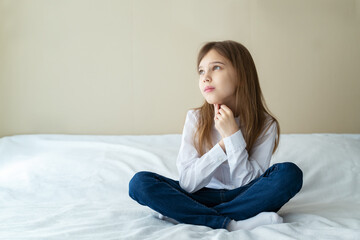 Naklejka premium Close up shot of beautiful blonde caucasian little girl wearing jeans and a white shirt, sitting on a bed and looking calm and dreamy