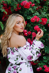 Naklejka premium Outdoor photo of romantic young woman in rose garden. girl with red lips in a dress with a print of roses. Young model on a background of a bush of roses. Stylish woman.