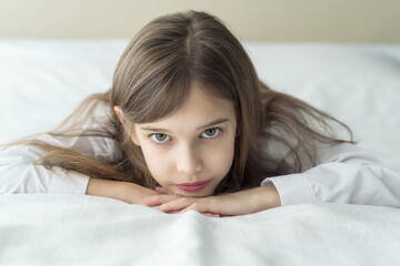 Close up shot of beautiful blonde caucasian little girl lying on a bed in a white shirt, looking calm and dreamy