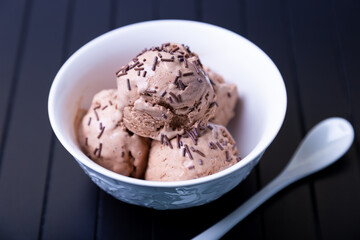 Chocolate ice cream with chocolate sprinkle in a white bowl on a black background. Three balls of homemade ice cream. Close-up.