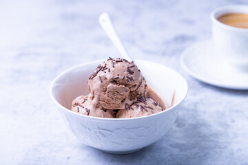 Chocolate ice cream with chocolate sprinkle in a white bowl on a light background. Three balls of homemade ice cream and a cup of coffee. Close-up.