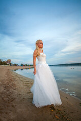 Fototapeta na wymiar Happy woman bride in long wedding white dress on sandy beach by the river on background of colorful sky
