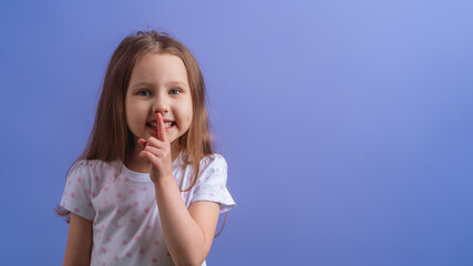 portrait of a cute little girl, holding her index finger to her mouth, stands on a purple background. The child doesn't want to talk. Keeps a secret, learned a secret