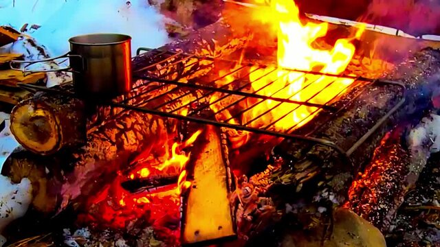 manmade log cabin style firepit with a metal gril on it boiling hot chocolate coffee steaming out with viger fire flaming with wooden plywood planks fed into the fire on a winter snow covered grounds