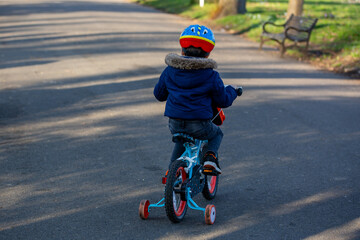 A small boy wearing a cycle helmet riding a bike in the park with stabilisers or stabilizers