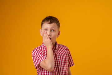 surprised boy, covering his mouth with his hand, stands on a yellow background. The child is upset, made a mistake. The guy is scared of the situation, he has a toothache.