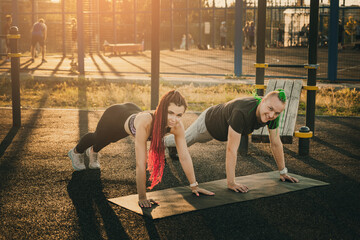 Fototapeta na wymiar A man and a woman with interesting hairstyles do a plank exercise on a city sports ground
