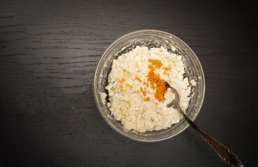 On a black table, orange zest is added to a glass bowl with mixed granular cottage cheese, egg, flour, close-up with copy space, top view