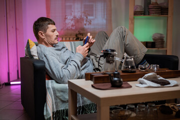 Home cozy atosphere on self-isolation. A young man is lying on the couch and talking on a video call with relatives. In front of him on the table is a set for the tea ceremony