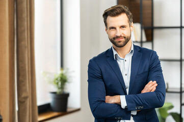 Successful confident young bearded brunet adult male entrepreneur businessman professional in business casual clothes standing with arms crossed folded at the modern home office and looking at camera - Powered by Adobe