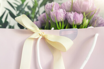 Close-up detail of fresh beautiful present of tender violet peony tulips bouquet and paper gift bag with romantic bow. Wedding or anniversary celebration surprise background. Pastel floral decoration