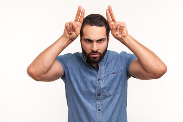 Naklejka premium Aggressive bully man with beard holding fingers above head showing horns, arrogant and stubborn, ready to attack, looking at camera and threatening. Indoor studio shot isolated on white background