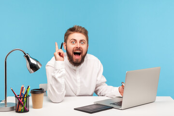 Smiling bearded man fashion designer pointing finger up, having idea, drawing with stylus on graphic tablet, sitting in office working on new collection. Indoor studio shot isolated on blue background