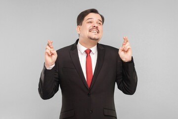 Businessman crosses his fingers and hopes for luck. indoor studio shot. isolated on gray background. handsome businessman with black suit, red tie and mustache looking at camera.
