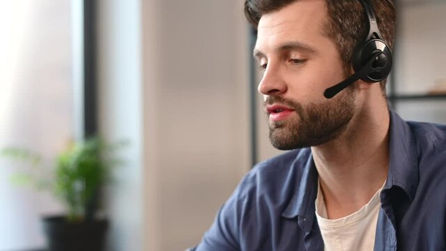 Smiling young bearded man wearing headset talking online, positive hipster in casual wear working in the customer service department, making and receiving calls, sitting in the home office, cropped