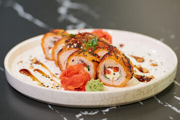 A close-up photo of sushi rolls with salmon, Philadelphia cheese, and tuna on a white ceramic plate with sauce, peanuts, ginger, and wasabi on a black stone table.