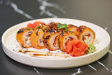 A photo of sushi rolls with salmon, Philadelphia cheese, and tuna on a white ceramic plate with sauce, peanuts, ginger, and wasabi on a black stone table.