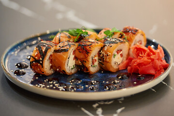 A close-up photo of fresh sushi rolls with salmon, Philadelphia cheese, and tuna on a blue ceramic plate with sauce, sesame,  lobules of kumquat, wasabi, and leaves. Black stone table.