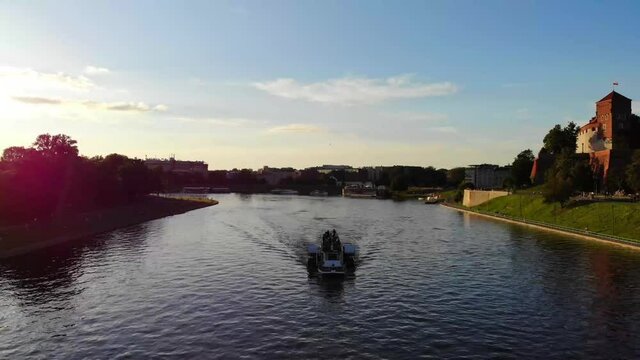 Drone shot of riverboat passing Wawel Castle on the right. The river is called the Vistula. Located in Krakow, Poland.