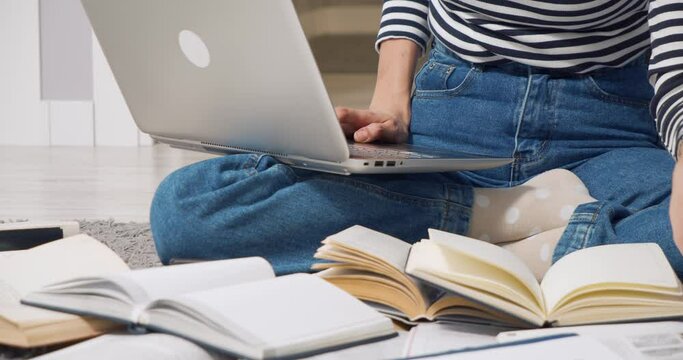 Unrecognizable young woman studying online or doing homework using laptop and books sitting on floor at home