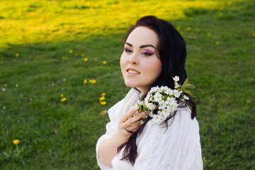 A girl sits on the grass in a park in a short white boho dress. The girl holds in her hands the first blossoms, a white branch of flowers. Brunette with long hair, spring has come. Gentle makeup
