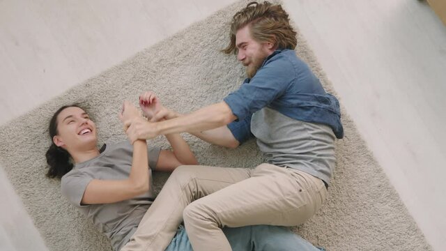 Top view shot of happy young man and woman lying on rug in their apartment and play fighting each other